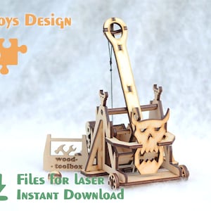 Puede incluir: Un modelo de madera de catapulta con un diseño de cara de demonio. El modelo tiene ruedas y una caja de herramientas. La imagen incluye el texto "Toys Design", "Files for Laser" e "Instant Download".