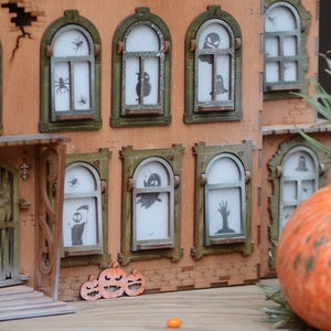 May include: A wooden model of a haunted house, perfect for Halloween, with arched windows displaying spooky silhouettes of spiders, ghosts, and a zombie hand. An orange pumpkin and carved pumpkin cutouts are in the foreground. The house is brown and green.