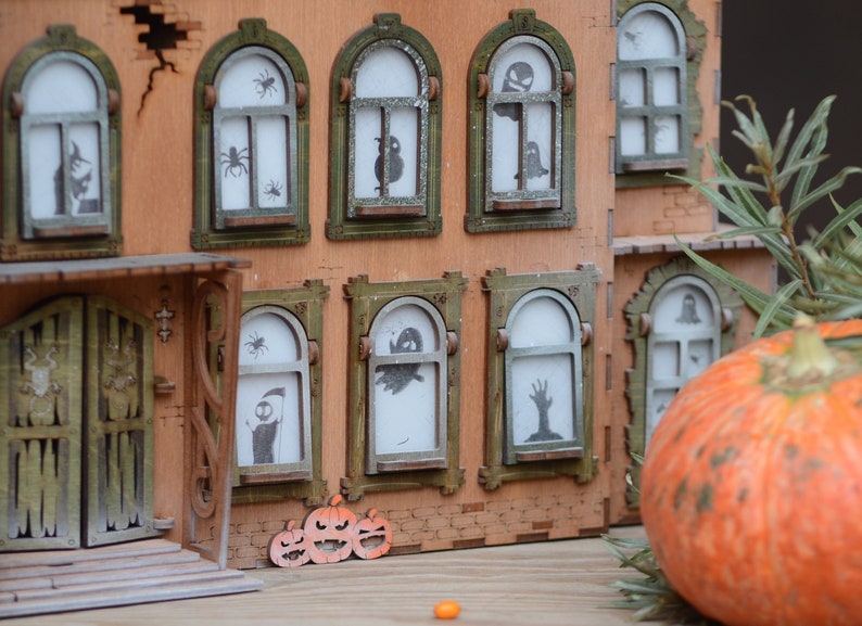 May include: A wooden model of a haunted house, perfect for Halloween, with arched windows displaying spooky silhouettes of spiders, ghosts, and a zombie hand. An orange pumpkin and carved pumpkin cutouts are in the foreground. The house is brown and green.