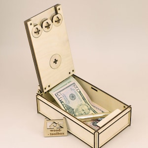 Op de afbeelding: Een lichtgekleurde houten kist met een scharnierend deksel, open om verschillende vijftig dollar biljetten en een pen te laten zien. De kist heeft een klein, apart houten bordje met de tekst "wood-toolbox". Het deksel heeft drie decoratieve schroefkoppen.
