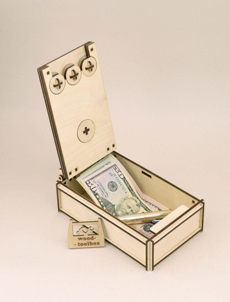 Op de afbeelding: Een lichtgekleurde houten kist met een scharnierend deksel, open om verschillende vijftig dollar biljetten en een pen te laten zien. De kist heeft een klein, apart houten bordje met de tekst "wood-toolbox". Het deksel heeft drie decoratieve schroefkoppen.