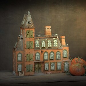 May include: A detailed orange and brown miniature haunted house with green shutters and a tall tower. The house has multiple windows with spooky silhouettes and is decorated with a bat and a small sign. Two pumpkins and greenery sit nearby.