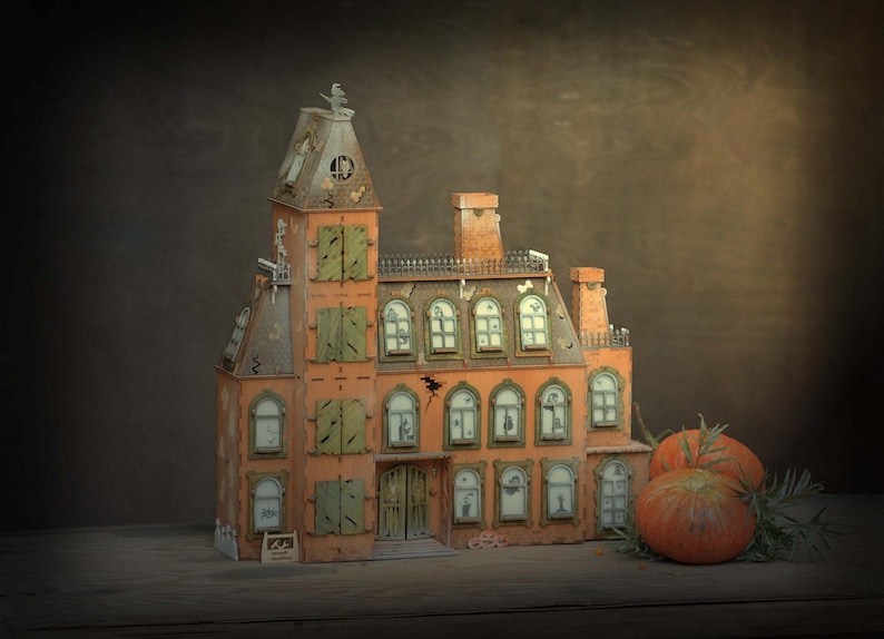 May include: A detailed orange and brown miniature haunted house with green shutters and a tall tower. The house has multiple windows with spooky silhouettes and is decorated with a bat and a small sign. Two pumpkins and greenery sit nearby.