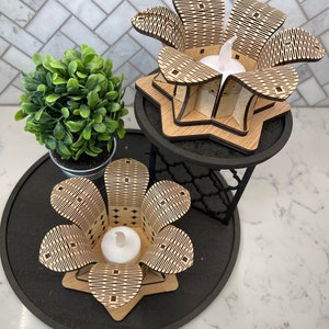 Puede incluir: Dos portavelas de madera con forma de flores con intrincados patrones geom&eacute;tricos. Los portavelas est&aacute;n sobre una bandeja negra de varios niveles con una peque&ntilde;a planta verde en una maceta.