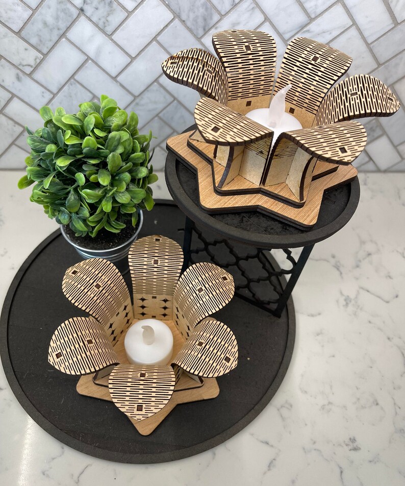 Puede incluir: Dos portavelas de madera con forma de flores con intrincados patrones geom&eacute;tricos. Los portavelas est&aacute;n sobre una bandeja negra de varios niveles con una peque&ntilde;a planta verde en una maceta.