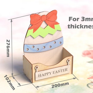 May include: Wooden Easter egg-shaped box with a bow, a blue and white stripe, and a green and yellow design. The box has a slot for holding items and a sign that says "Happy Easter". The box is 276 mm tall, 200 mm wide, and 102 mm deep.