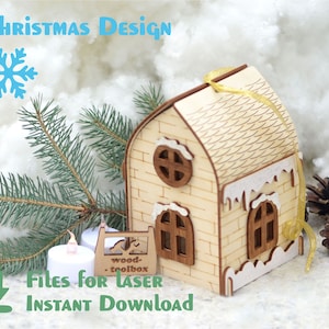 Op de afbeelding: Een houten kerst ornament in de vorm van een huis met een wit besneeuwd dak en een gouden lint. Het huis heeft een bakstenen gevel en twee ramen. Het ornament ligt op een wit, pluche oppervlak met een dennentak en dennenappels.