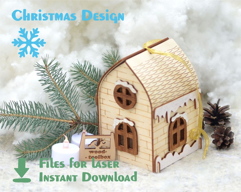Op de afbeelding: Een houten kerst ornament in de vorm van een huis met een wit besneeuwd dak en een gouden lint. Het huis heeft een bakstenen gevel en twee ramen. Het ornament ligt op een wit, pluche oppervlak met een dennentak en dennenappels.