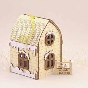 Op de afbeelding: Een houten kerstversiering in de vorm van een huis met een wit dak en sneeuw bedekte rand. Het huis heeft twee ramen en een rond raam aan de voorkant. De versiering hangt aan een geel lint en heeft de tekst "wood-toolbox" aan de onderkant.