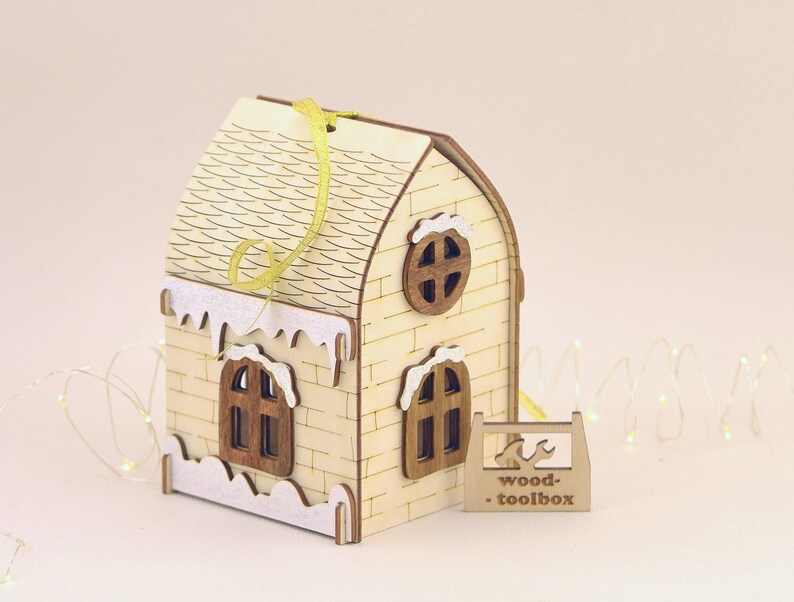 Op de afbeelding: Een houten kerstversiering in de vorm van een huis met een wit dak en sneeuw bedekte rand. Het huis heeft twee ramen en een rond raam aan de voorkant. De versiering hangt aan een geel lint en heeft de tekst "wood-toolbox" aan de onderkant.