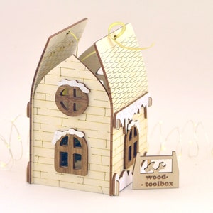 Op de afbeelding: Een houten huis ornament met een witte bakstenen gevel, een met sneeuw bedekt dak en een klein raam. Het huis is aan &eacute;&eacute;n kant open, waardoor een klein interieur zichtbaar is. Het ornament heeft een geel lint dat aan de bovenkant is bevestigd en een bord met de tekst "wood-toolbox".