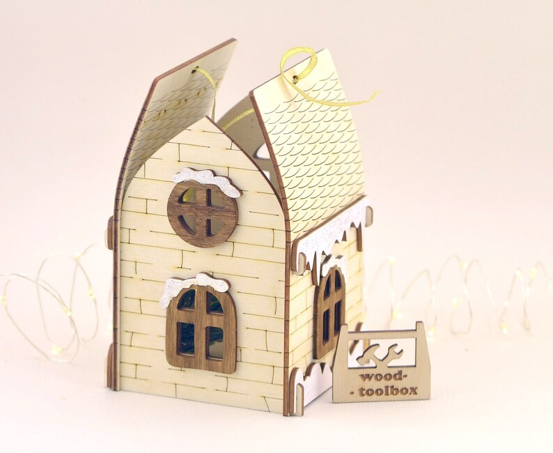 Op de afbeelding: Een houten huis ornament met een witte bakstenen gevel, een met sneeuw bedekt dak en een klein raam. Het huis is aan &eacute;&eacute;n kant open, waardoor een klein interieur zichtbaar is. Het ornament heeft een geel lint dat aan de bovenkant is bevestigd en een bord met de tekst "wood-toolbox".