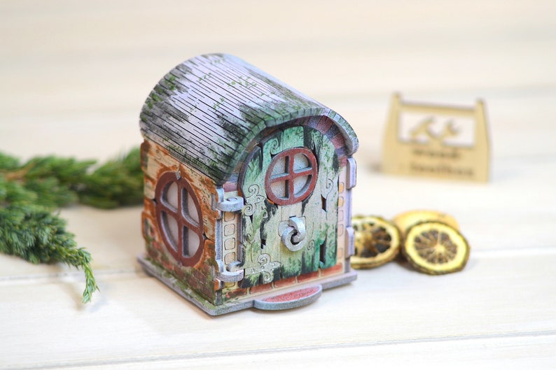 May include: A small wooden house with a green door and a window. The house is painted in shades of brown, green, and white, and has a weathered look. The house is sitting on a white surface.