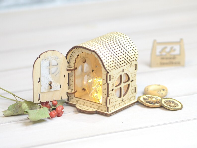 May include: A wooden house shaped nightlight with a door that opens and a window with a string of lights inside. The house is sitting on a white surface.
