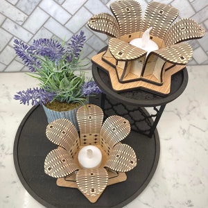 Puede incluir: Dos portavelas de madera con forma de flor con un patr&oacute;n geom&eacute;trico. Los portavelas est&aacute;n en una bandeja de niveles negros con una planta en maceta.