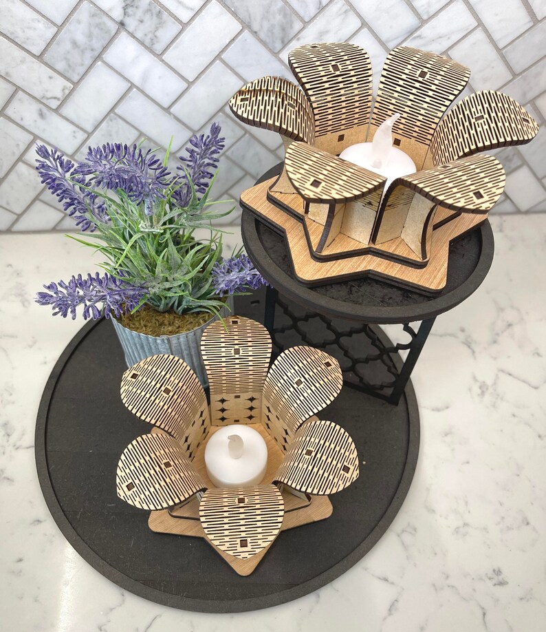 Puede incluir: Dos portavelas de madera con forma de flor con un patr&oacute;n geom&eacute;trico. Los portavelas est&aacute;n en una bandeja de niveles negros con una planta en maceta.