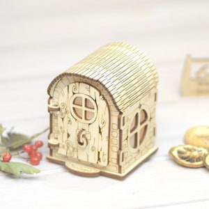 May include: A wooden fairy house with a light brown roof, a door with a window, and a small window on the side. The house is sitting on a white surface.