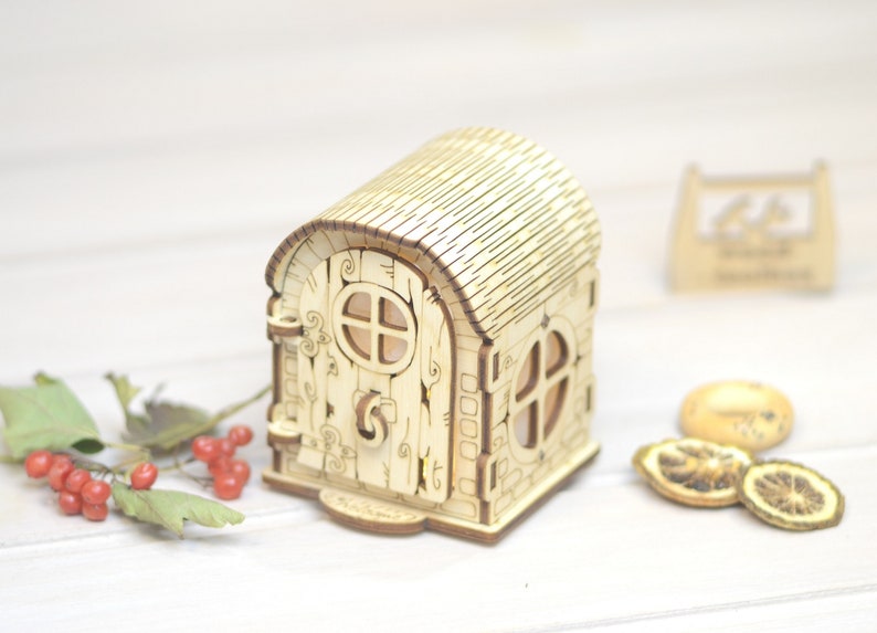 May include: A wooden fairy house with a light brown roof, a door with a window, and a small window on the side. The house is sitting on a white surface.