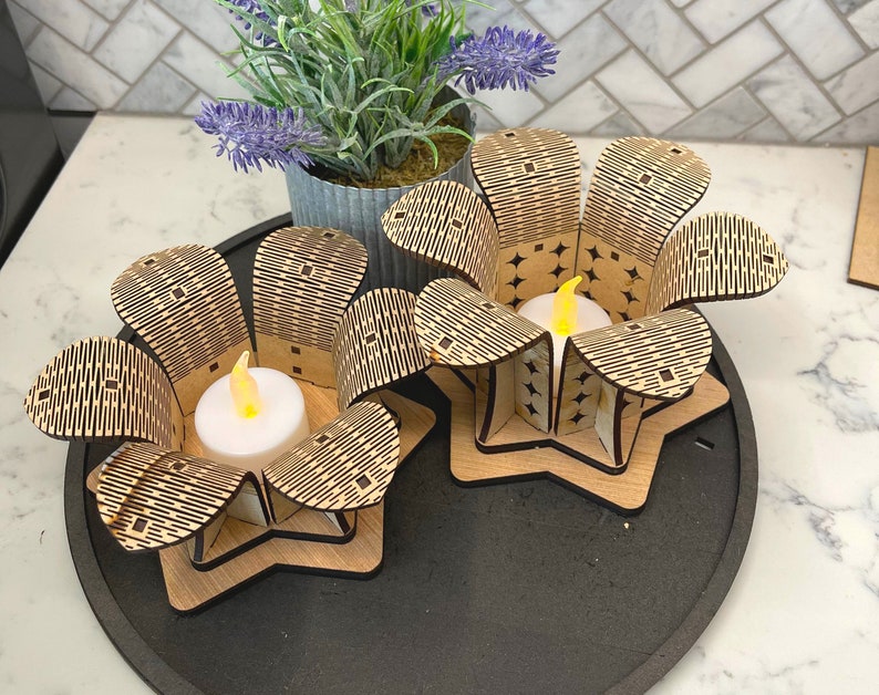 Puede incluir: Dos portavelas de madera con forma de flores con un patr&oacute;n geom&eacute;trico. Cada portavelas tiene una peque&ntilde;a vela de t&eacute; blanca en su interior.