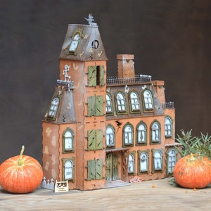 May include: A detailed, orange and brown haunted house model with green shutters, a witch on the roof, and ghost and bat cutouts. Two orange pumpkins sit in front of the house, creating a Halloween theme.