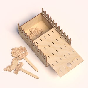 May include: A wooden garden box with a sliding lid and decorative fence-like edges. The box contains wooden plant markers labeled "Basil" and "Tomato". Additional markers for "Spinach" and "Eggplant" are placed nearby.