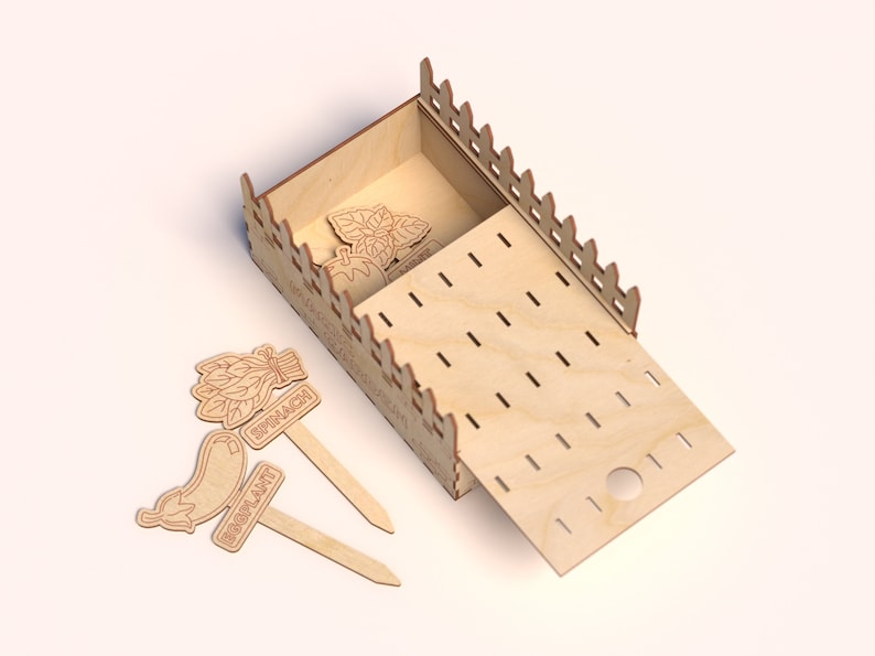 May include: A wooden garden box with a sliding lid and decorative fence-like edges. The box contains wooden plant markers labeled "Basil" and "Tomato". Additional markers for "Spinach" and "Eggplant" are placed nearby.