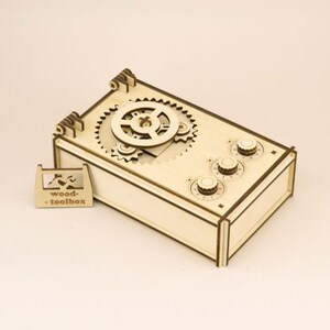 Op de afbeelding: Een houten puzzelbox met zichtbare tandwielen en wijzerplaten. De box is rechthoekig. De bovenkant heeft in elkaar grijpende tandwielen en drie ronde wijzerplaten. Een klein houten label met de tekst "wood-toolbox" staat naast de box. Afmetingen in centimeters.