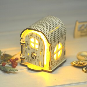 May include: A wooden house-shaped nightlight with a glowing window and a detailed door. The house is made of light wood and has a textured roof.