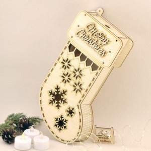 May include: A wooden Christmas stocking with a cutout design of snowflakes and the words "Merry Christmas". The stocking is light brown and has a small wooden stand. There are three white tea lights and a string of white fairy lights in the image.