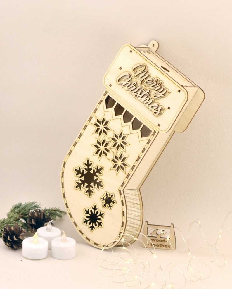 May include: A wooden Christmas stocking with a cutout design of snowflakes and the words "Merry Christmas". The stocking is light brown and has a small wooden stand. There are three white tea lights and a string of white fairy lights in the image.