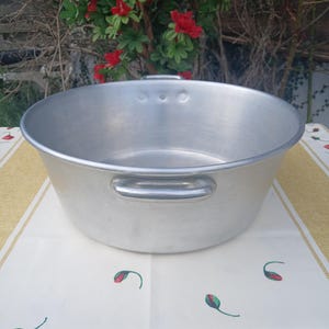 Peut inclure: Un grand bassin rond en aluminium avec deux poignées latérales. Le bassin a un large bord et une finition en métal brossé. Il repose sur une nappe à motifs floraux. L'arrière-plan présente des fleurs rouges et du feuillage vert.