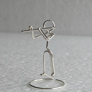 Op de afbeelding: Een zilveren draadsculptuur van een persoon die fluit speelt. De minimalistische figuur is gemaakt van één stuk draad en toont een muzikant met een fluit, staand op een cirkelvormige basis. Het beeld is een decoratief item.