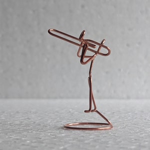 May include: A copper wire sculpture of a person playing a trombone. The musician is standing on a circular base. The trombone is held up to the musician's face. The sculpture is made of a single piece of copper wire.