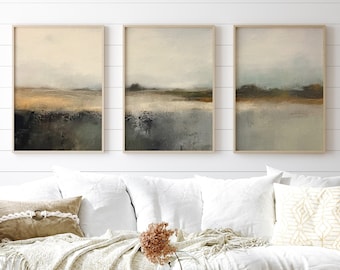 May include: Three abstract landscape paintings in muted tones of brown, green, and blue, hanging on a white wall above a white couch with beige pillows and a throw blanket. A vase with pink flowers sits on a wooden coffee table in front of the couch.