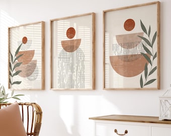 May include: Three framed abstract art prints with geometric shapes and foliage in shades of brown, orange, and green. The prints are hung on a white wall above a white cabinet with a wood top.