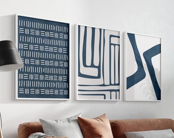 May include: Three abstract art prints in white frames with a brown leather sofa, a woven chair, and a potted plant in the background. The prints feature geometric shapes in shades of blue and white.