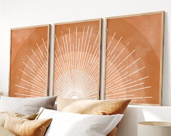 May include: Three orange and white abstract sunburst prints with a wood frame hanging above a bed with tan bedding and pillows.