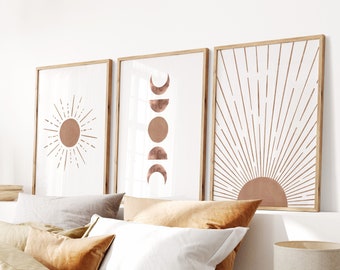 May include: Three minimalist wall art prints with a white background and copper colored sun, moon, and sunburst designs. The prints are framed in natural wood and are displayed above a bed with a tan duvet cover and pillows.