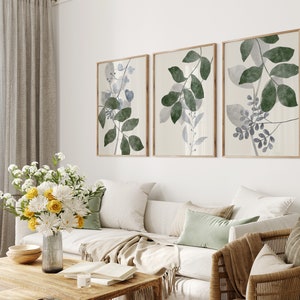 K&ouml;nnte beinhalten: Drei gerahmte botanische Drucke mit einem hellbeigen Hintergrund und gr&uuml;nen und blauen Aquarellbl&auml;ttern. Die Drucke h&auml;ngen an einer wei&szlig;en Wand &uuml;ber einem wei&szlig;en Sofa mit beigen Kissen und einer braunen Wolldecke.