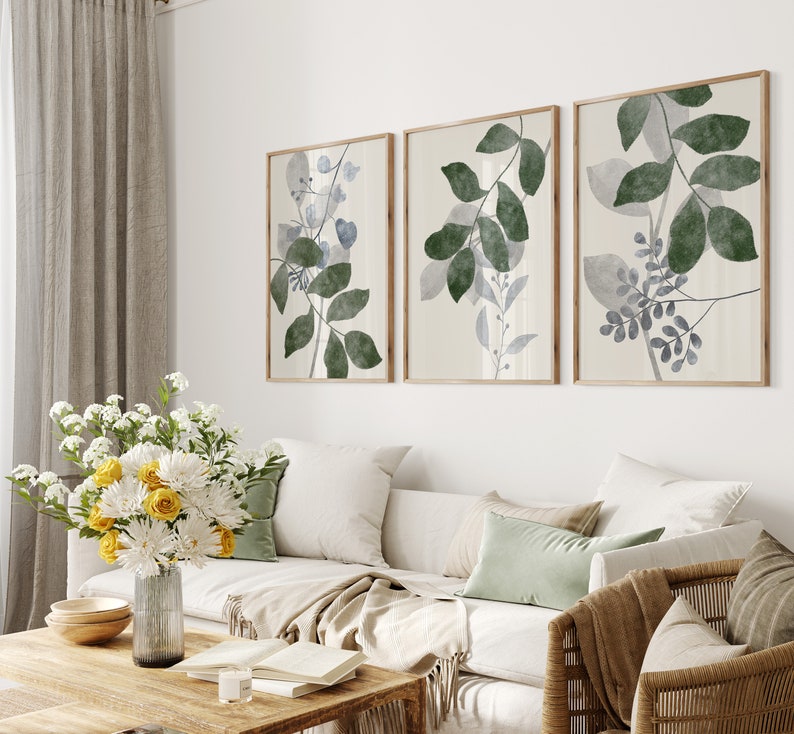 K&ouml;nnte beinhalten: Drei gerahmte botanische Drucke mit einem hellbeigen Hintergrund und gr&uuml;nen und blauen Aquarellbl&auml;ttern. Die Drucke h&auml;ngen an einer wei&szlig;en Wand &uuml;ber einem wei&szlig;en Sofa mit beigen Kissen und einer braunen Wolldecke.