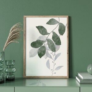 K&ouml;nnte beinhalten: Ein gerahmter Druck eines Aquarells mit gr&uuml;nen und grauen Bl&auml;ttern auf wei&szlig;em Hintergrund. Der Druck h&auml;ngt an einer gr&uuml;nen Wand &uuml;ber einer Kommode mit zwei gr&uuml;nen Glasvasen und einem Stapel B&uuml;chern mit einer gr&uuml;nen Kugel darauf.