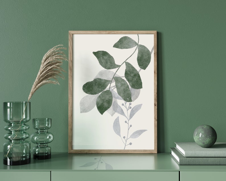 K&ouml;nnte beinhalten: Ein gerahmter Druck eines Aquarells mit gr&uuml;nen und grauen Bl&auml;ttern auf wei&szlig;em Hintergrund. Der Druck h&auml;ngt an einer gr&uuml;nen Wand &uuml;ber einer Kommode mit zwei gr&uuml;nen Glasvasen und einem Stapel B&uuml;chern mit einer gr&uuml;nen Kugel darauf.