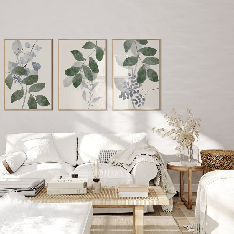K&ouml;nnte beinhalten: Drei gerahmte Aquarelldrucke von stilisierten gr&uuml;nen und blauen Bl&auml;ttern an einer wei&szlig;en Wand &uuml;ber einem wei&szlig;en Sofa mit einer Wolldecke und einem Couchtisch mit B&uuml;chern und einem Diffusor.
