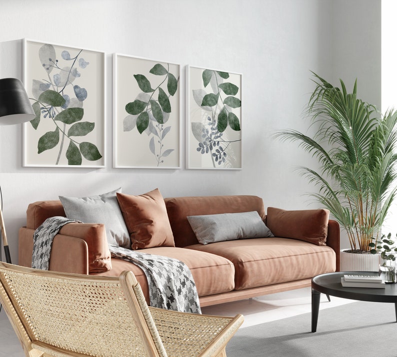 K&ouml;nnte beinhalten: Drei gerahmte botanische Drucke mit einem hellbeigen Hintergrund und gr&uuml;nen und blauen Aquarellbl&auml;ttern. Die Drucke h&auml;ngen an einer wei&szlig;en Wand &uuml;ber einem braunen Sofa mit zwei Kissen und einer Wolldecke.