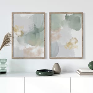 Op de afbeelding: Twee ingelijste abstracte kunstprints met een aquarelontwerp in tinten groen, grijs en goud. De prints hangen aan een witte muur boven een witte commode.