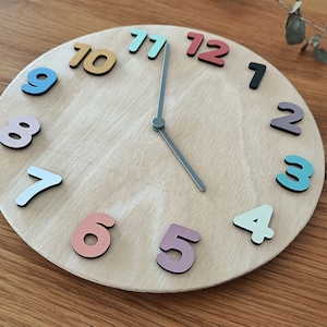 Könnte beinhalten: Eine runde Holzuhr mit bunten Zahlen. Die Zahlen sind in verschiedenen Farben, darunter Blau, Rot und Gelb. Die Uhrzeiger sind grau. Die Uhr befindet sich auf einer Holzoberfläche.