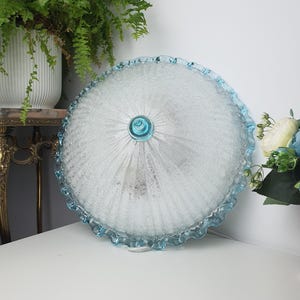 May include: A round, dome-shaped glass ceiling light fitting featuring a textured, frosted surface with radiating ribs. The edges are finished with a ruffled, light blue glass trim, and a small, matching blue glass finial sits at the centre.