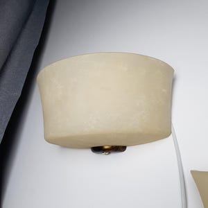 May include: A semi-circular wall sconce featuring a frosted, off-white glass shade with a mottled texture. The fixture is secured to the wall with a dark, bronze-toned metal base and includes a white power cord extending from the bottom.