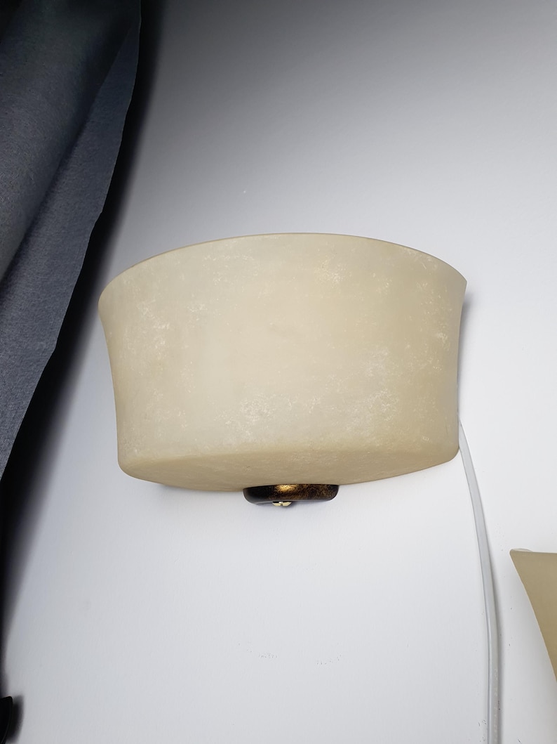 May include: A semi-circular wall sconce featuring a frosted, off-white glass shade with a mottled texture. The fixture is secured to the wall with a dark, bronze-toned metal base and includes a white power cord extending from the bottom.