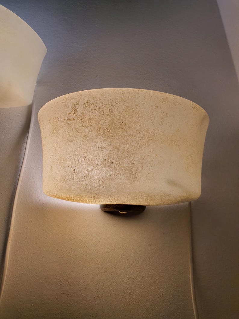 May include: A semi-circular wall sconce featuring a textured, off-white, translucent glass shade with mottled brown and tan speckling. The shade is mounted to the wall with a dark, circular base, emitting a soft glow from the bottom edge.